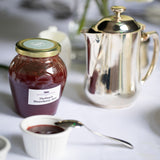 A jar of Thelma's homemade strawberry jam is placed on a table, with a silver teapot and a spoon with jam on it in the foreground.